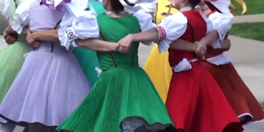 Groep mensen in traditionele kleurrijke klederdracht dansen in een kring, met de armen om elkaars schouders geslagen. De kleding bestaat uit wijde rokken en geborduurde blouses.
