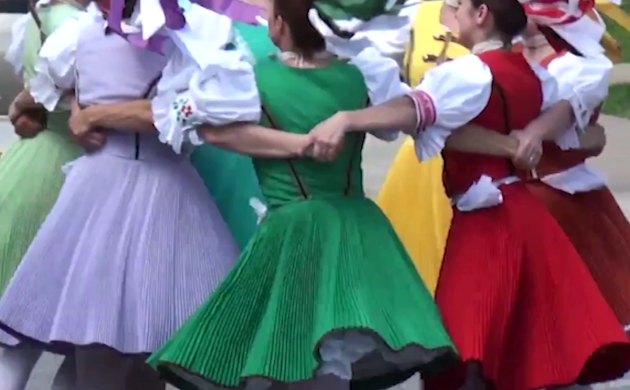 Groep mensen in traditionele kleurrijke klederdracht dansen in een kring, met de armen om elkaars schouders geslagen. De kleding bestaat uit wijde rokken en geborduurde blouses.