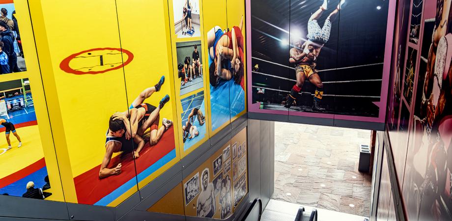 Grote blauwe wandpanelen tonen foto's van boksers. Mannen en vrouwen trainen in de boksring. Eén bokser rust uit op de touwen.